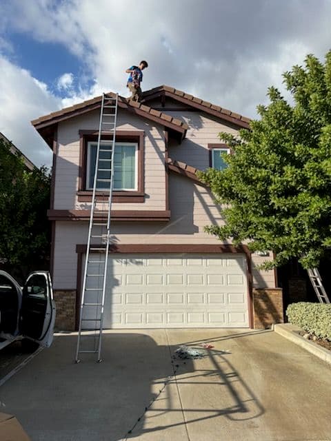 Child on ladder cleaning roof of a pink house with garage, surrounded by trees and blue sky.