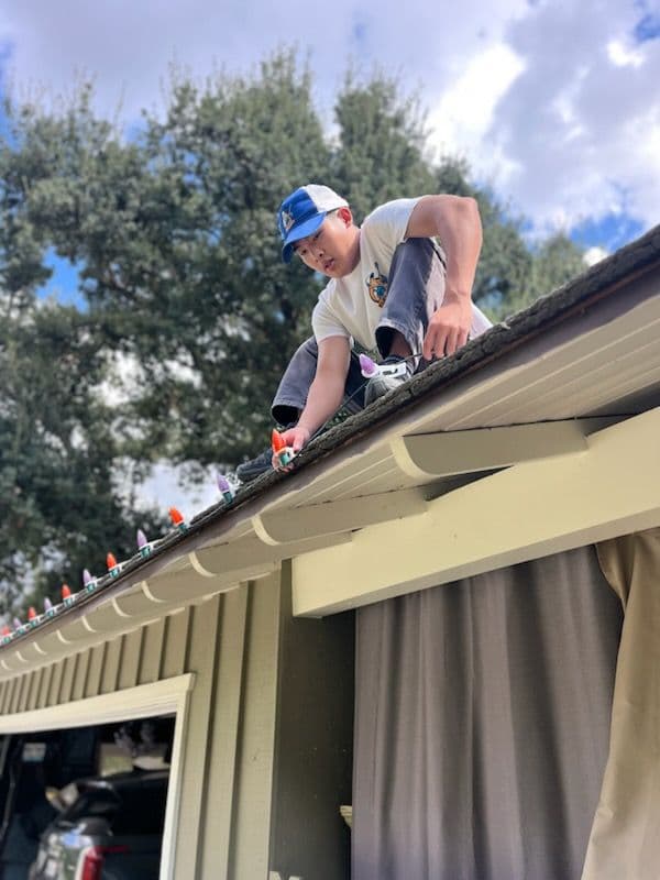 Person installing holiday decorations on a roof with colorful lights, against a cloudy sky.