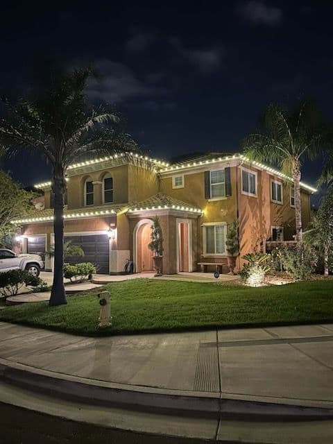Two-story house with palm trees, illuminated by festive lights at night. Green lawn in front.
